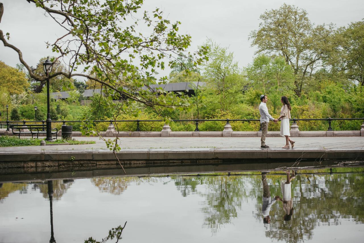 Wide engagement portrait of a couple beside a reflecting pool beneath spring trees