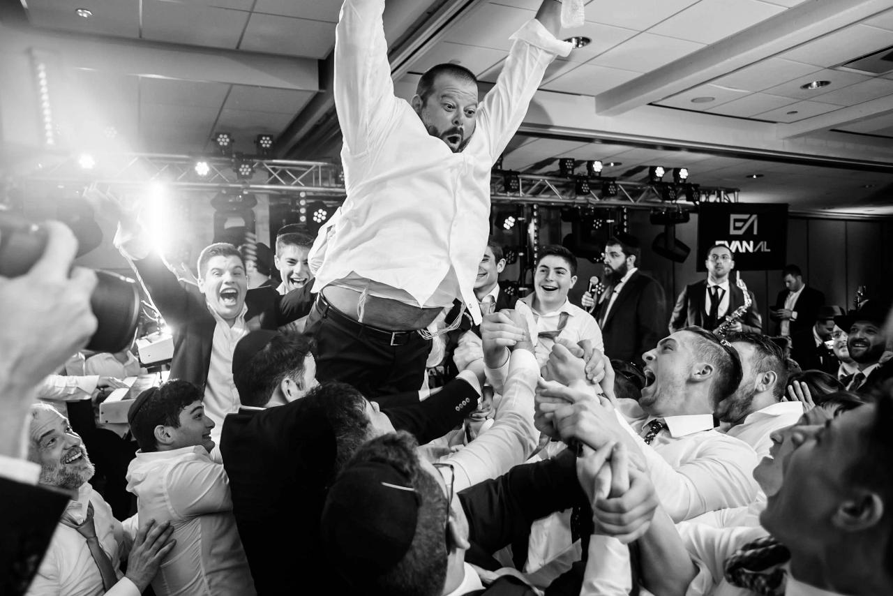 Black-and-white wedding reception dance moment with a man lifted high overhead by the crowd as guests smile and cheer in a banquet hall.