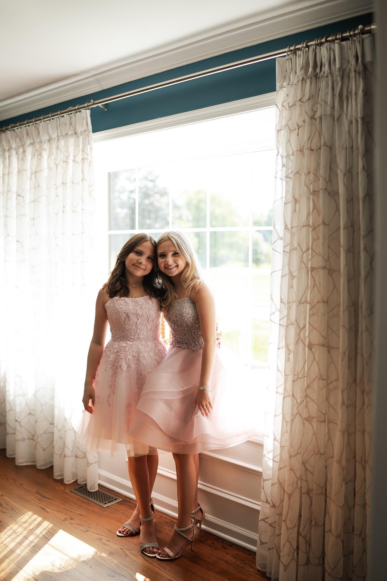 Twin sisters in pink dresses standing together beside a sunlit window
