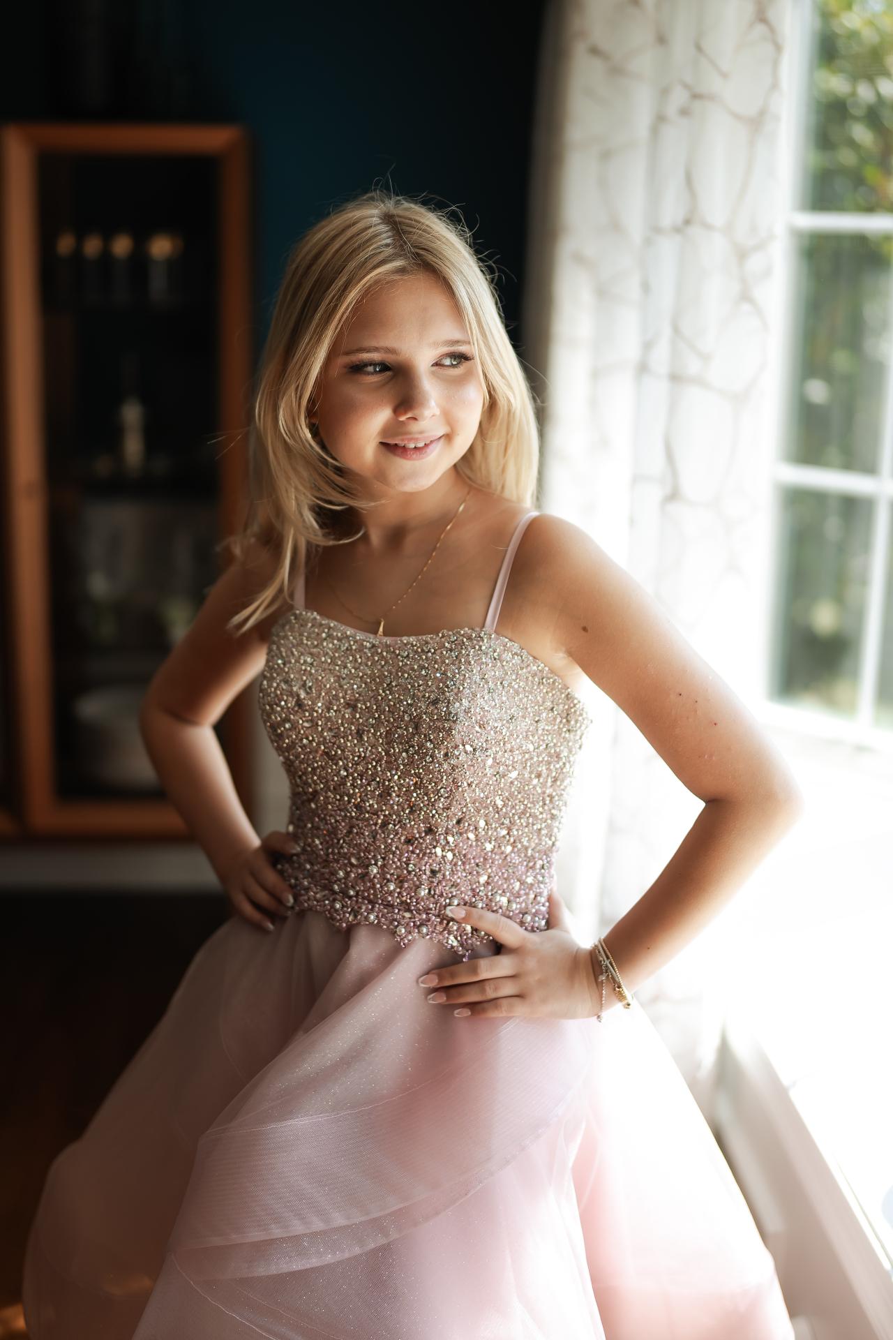 Bat mitzvah portrait of a sister smiling beside a window in a pink dress