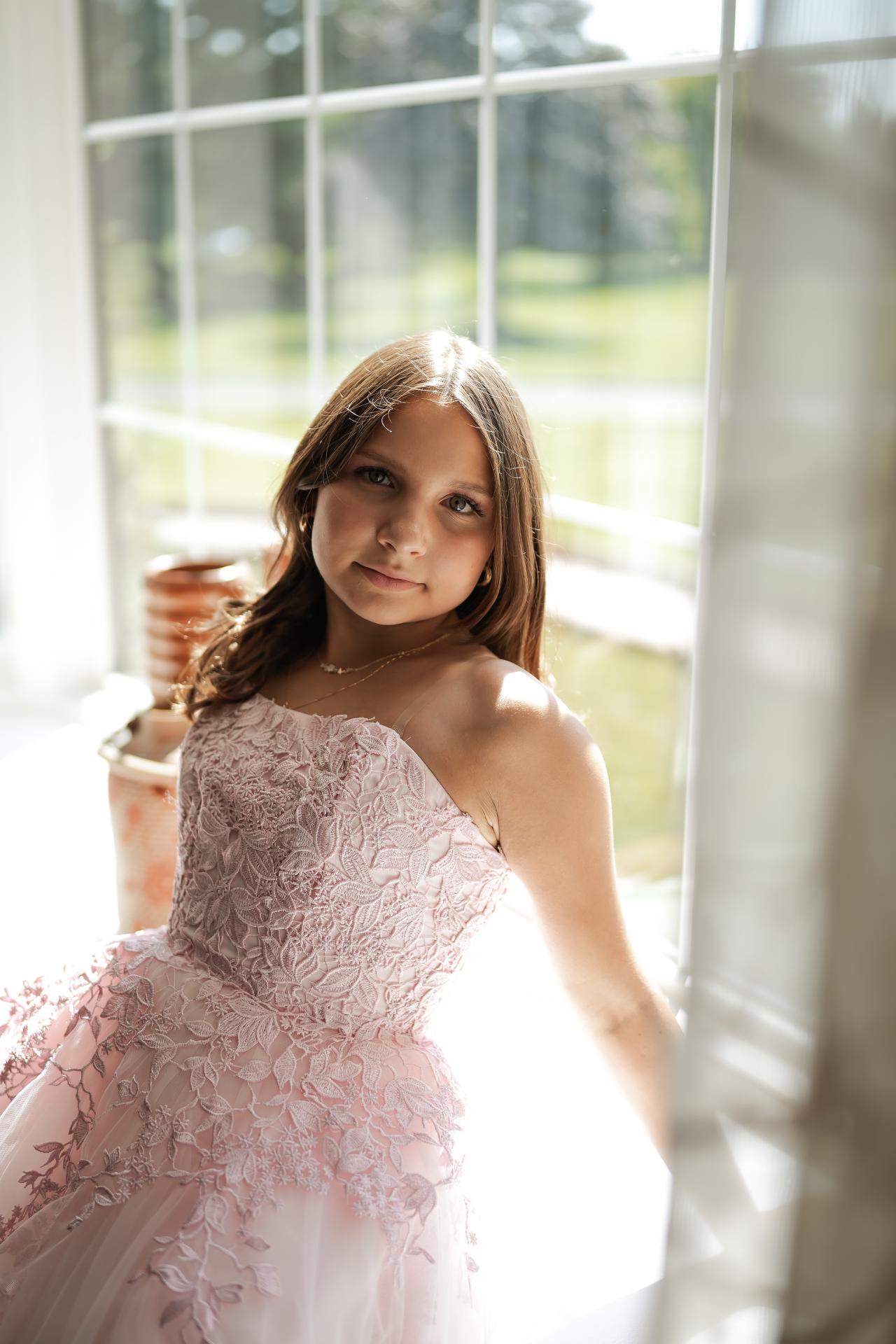 Portrait of a sister in a pink lace dress beside a bright window