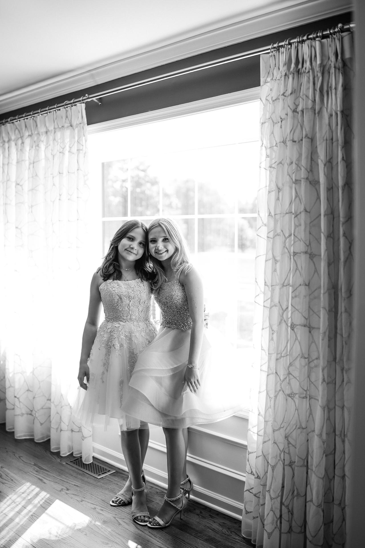 Black-and-white portrait of twin sisters standing together beside a window