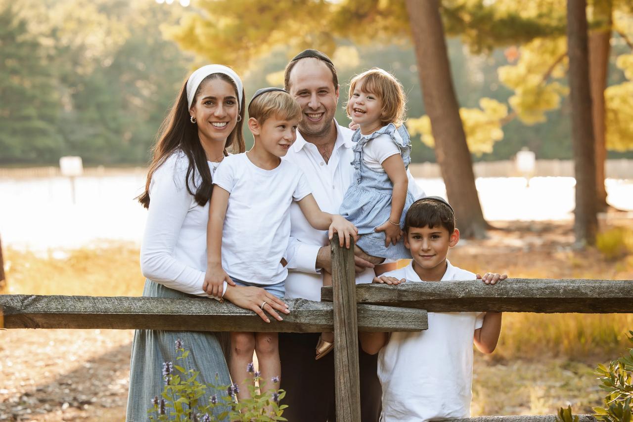 Family of five smiling beside a wooden fence and autumn trees by the water