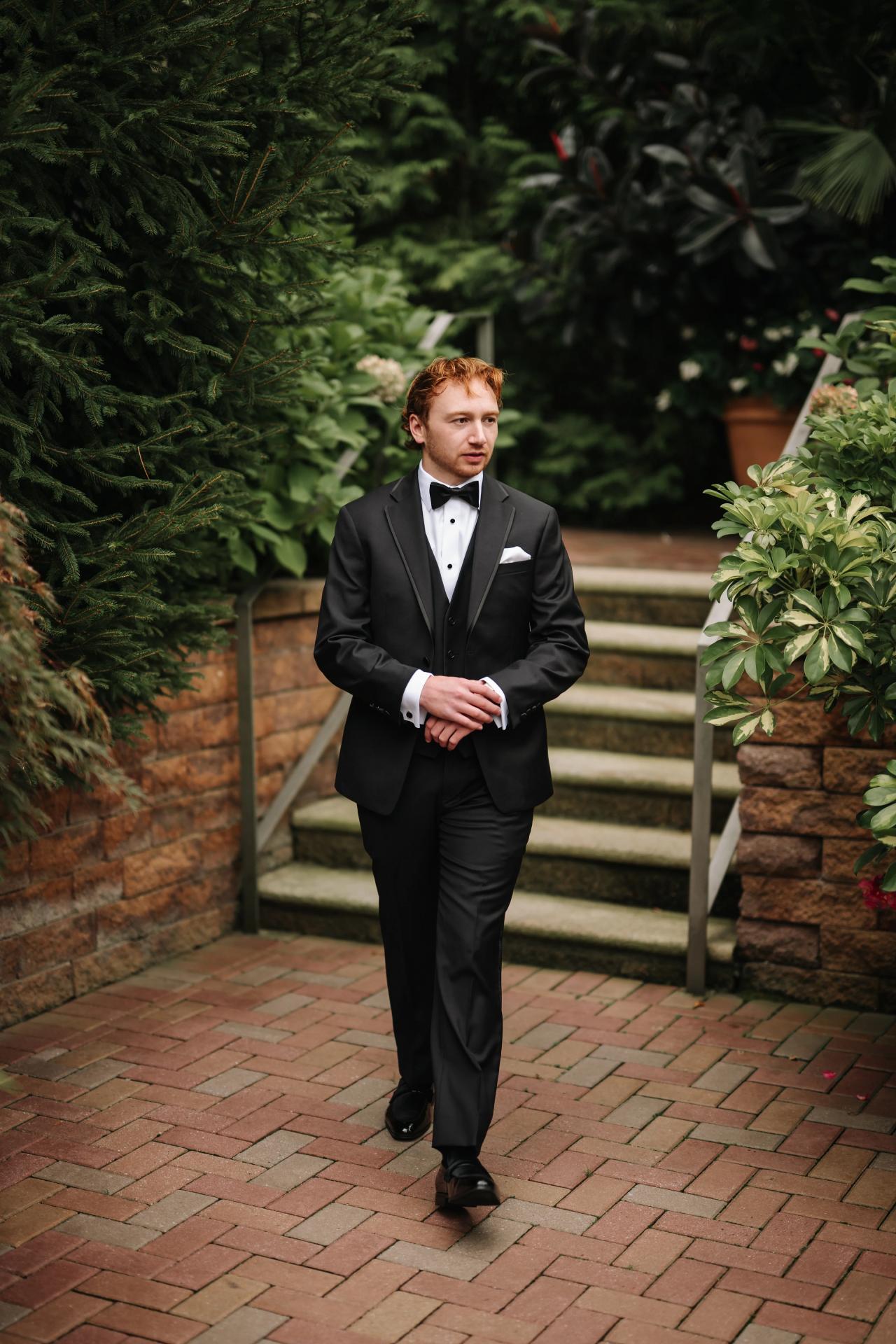Groom standing in a tuxedo beneath garden greenery. Crest Hollow wedding in Woodbury, Long Island.