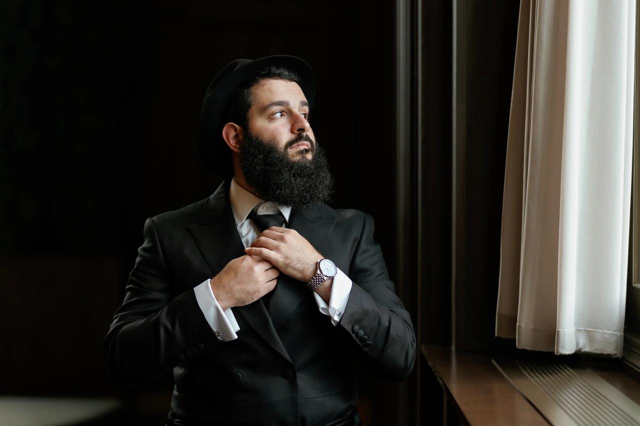Groom adjusting his tie beside a sunlit window. Jewish wedding in Detroit.