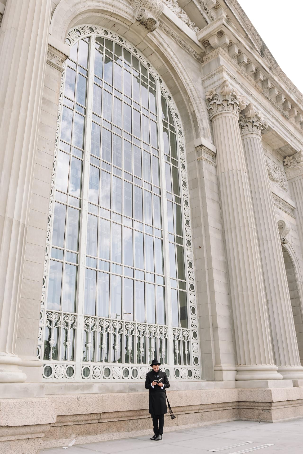 Wide groom portrait beneath monumental windows and stone columns. Jewish wedding in Detroit.