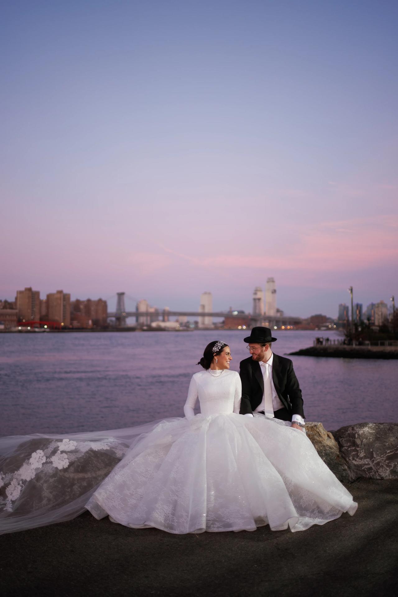 Bride and groom seated by the river beneath a pastel sky. Wedding portraits in Brooklyn, NYC.