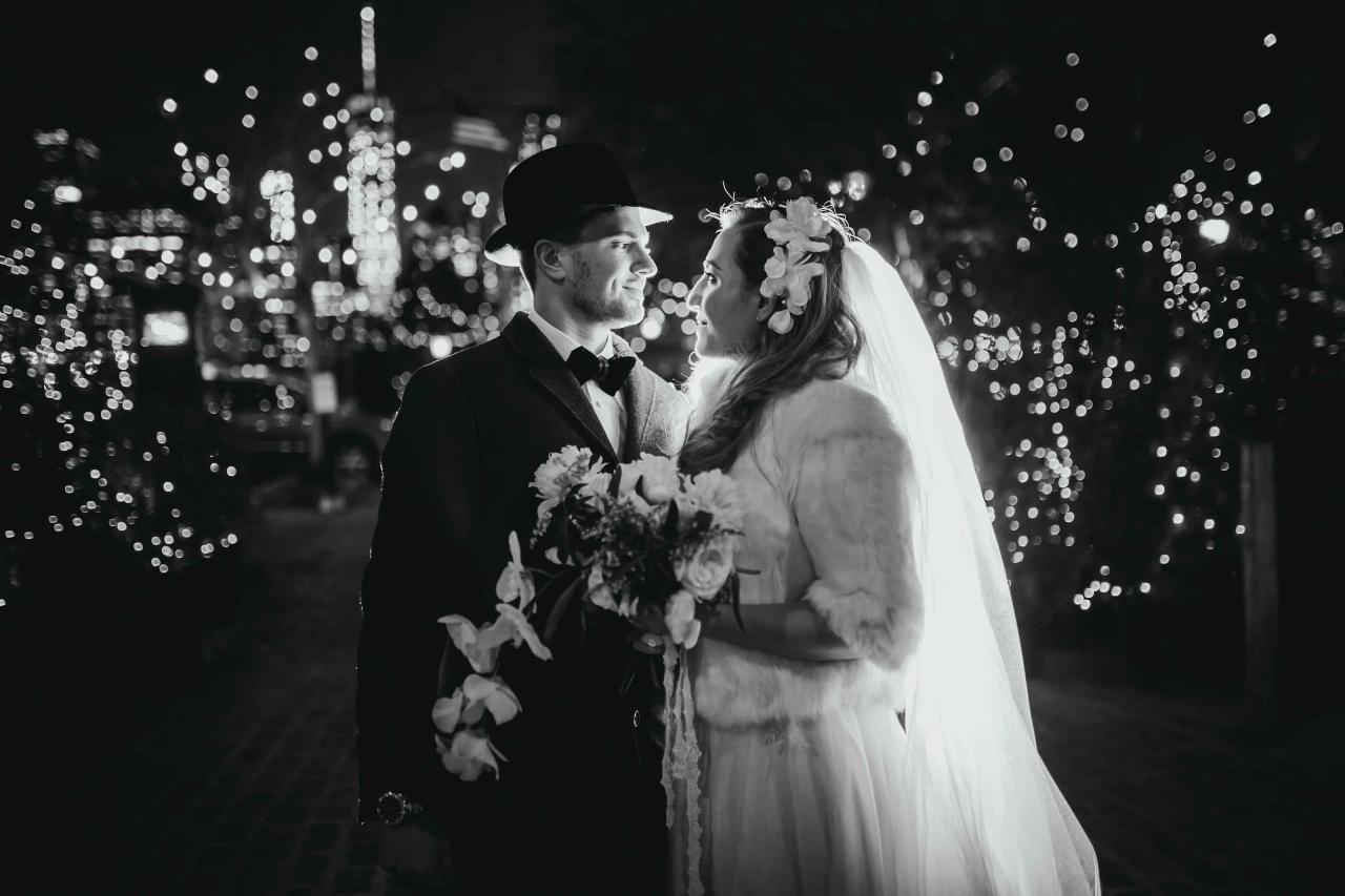 Black-and-white wedding portrait of a bride and groom under night lights, holding bouquets and looking at each other while standing close together.