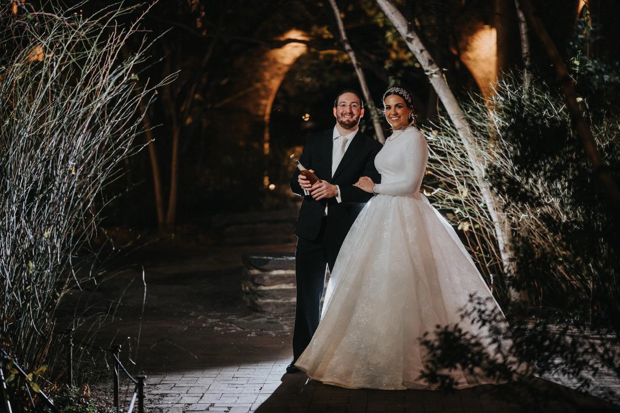Bride and groom opening a bottle beneath twinkling garden lights. Wedding portraits in Brooklyn, NYC.