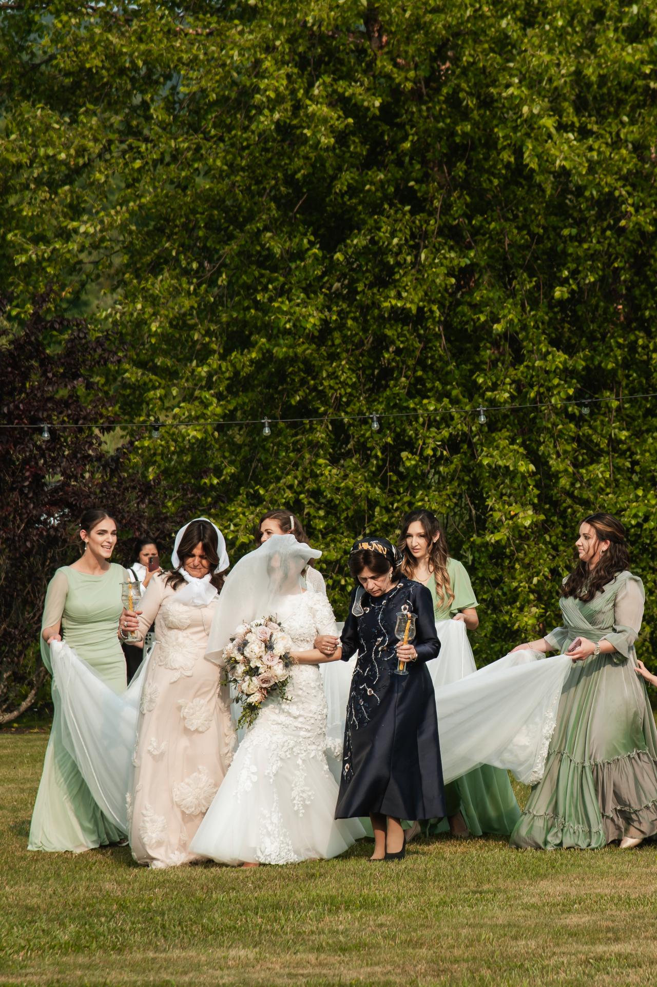 Bride approaching the chuppah with family beneath summer trees. Wedding at Indian Ridge.