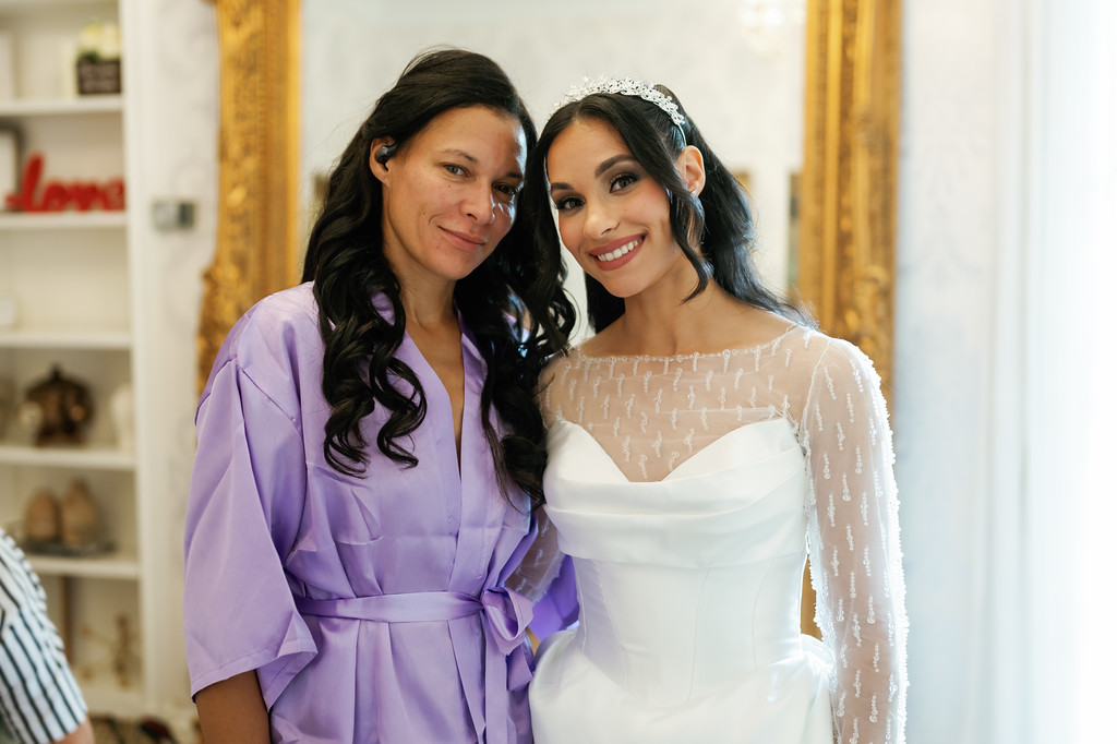 A smiling bride in a white dress stands close with a woman in a lavender robe indoors, both looking toward the camera.