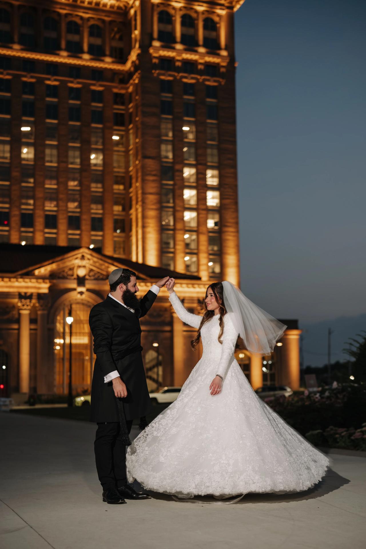 Groom twirling the bride before a warmly illuminated Detroit facade at dusk