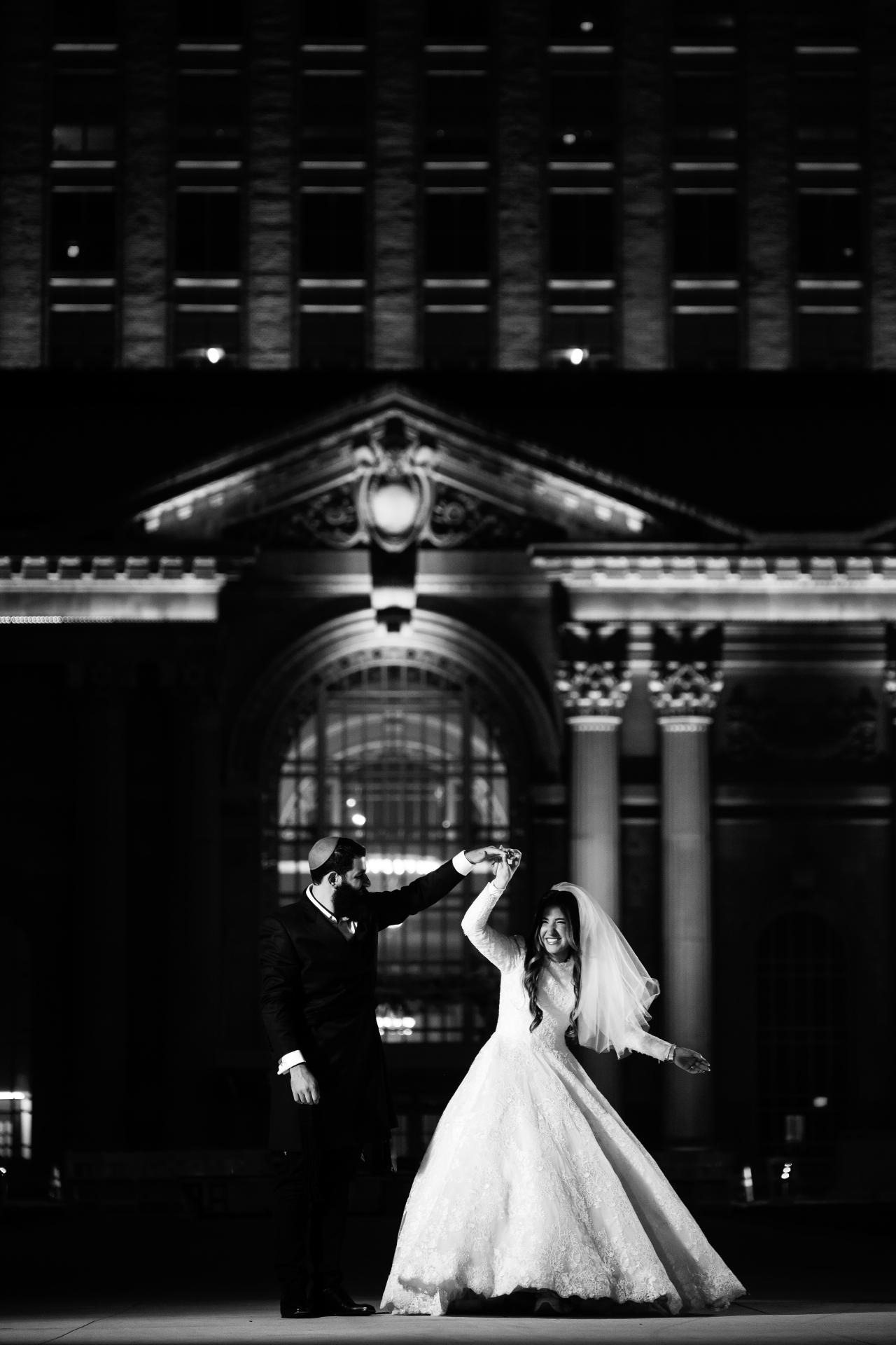 Black-and-white groom twirling the bride beneath illuminated stone arches at night in Detroit