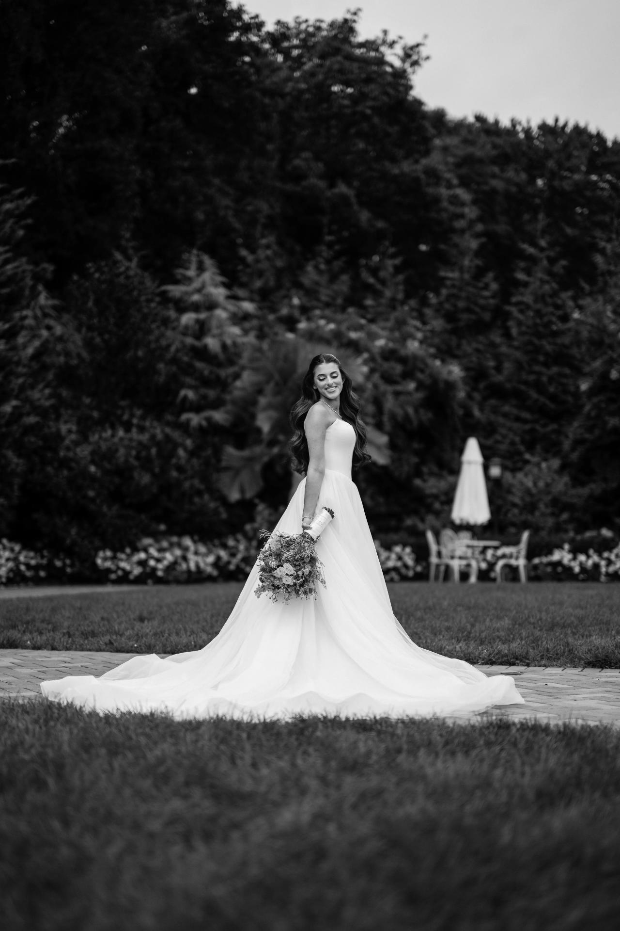 Black-and-white bride with a long train against dark gardens and soft foreground grass at Crest Hollow, Woodbury, Long Island.