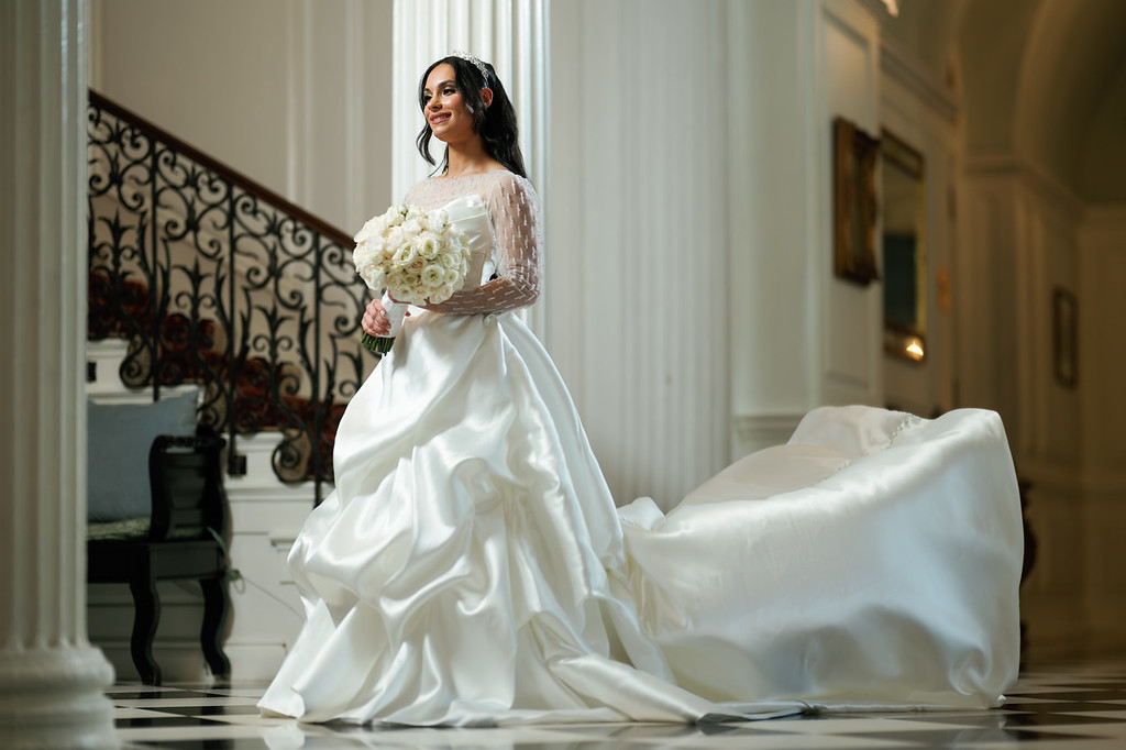 Bride in a white wedding dress holding a bouquet while her long train is spread on the floor, framed by an elegant staircase and doorway.