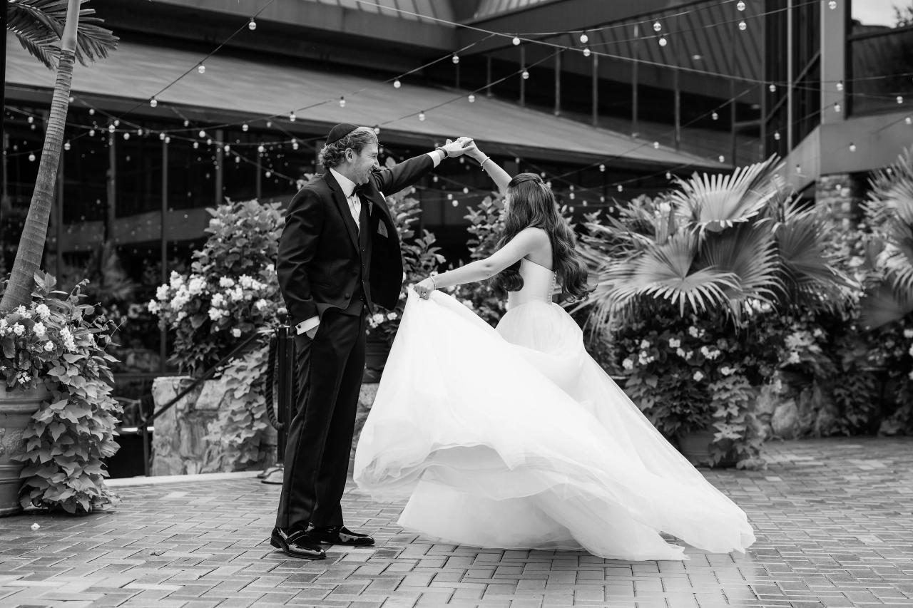 Black-and-white groom twirling the bride beneath string lights in the Crest Hollow gardens, Woodbury, Long Island.