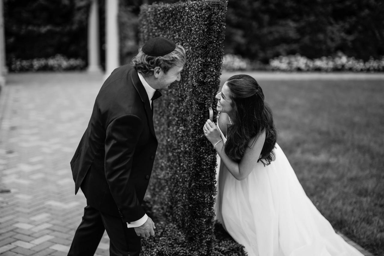 Black-and-white bride and groom leaning toward each other around a tall garden topiary at Crest Hollow, Woodbury, Long Island.