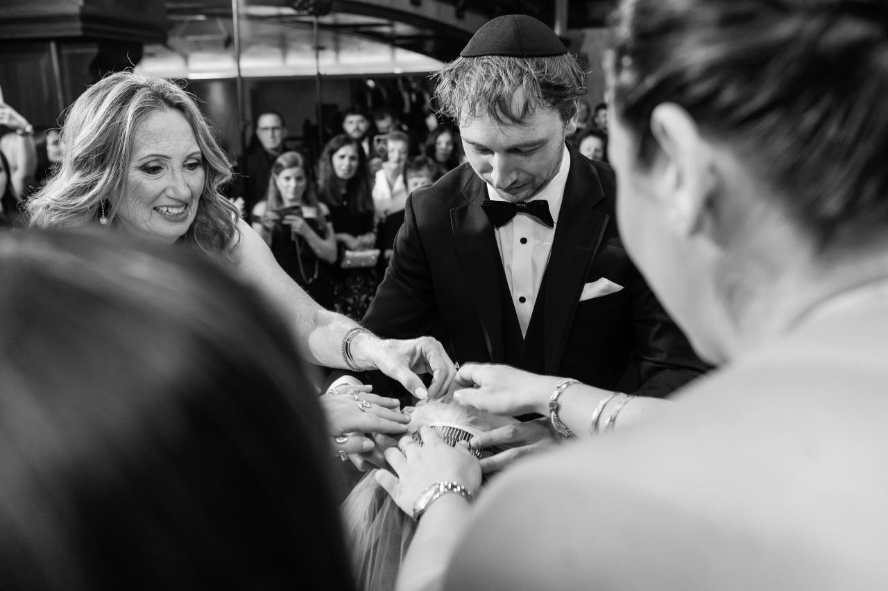 Black-and-white groom helping fasten the bridal veil, framed by softly blurred family members at Crest Hollow, Woodbury, Long Island.
