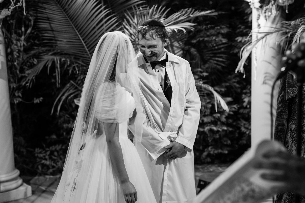 Black-and-white smiling groom looking toward his veiled bride beneath palm fronds at Crest Hollow, Woodbury, Long Island.