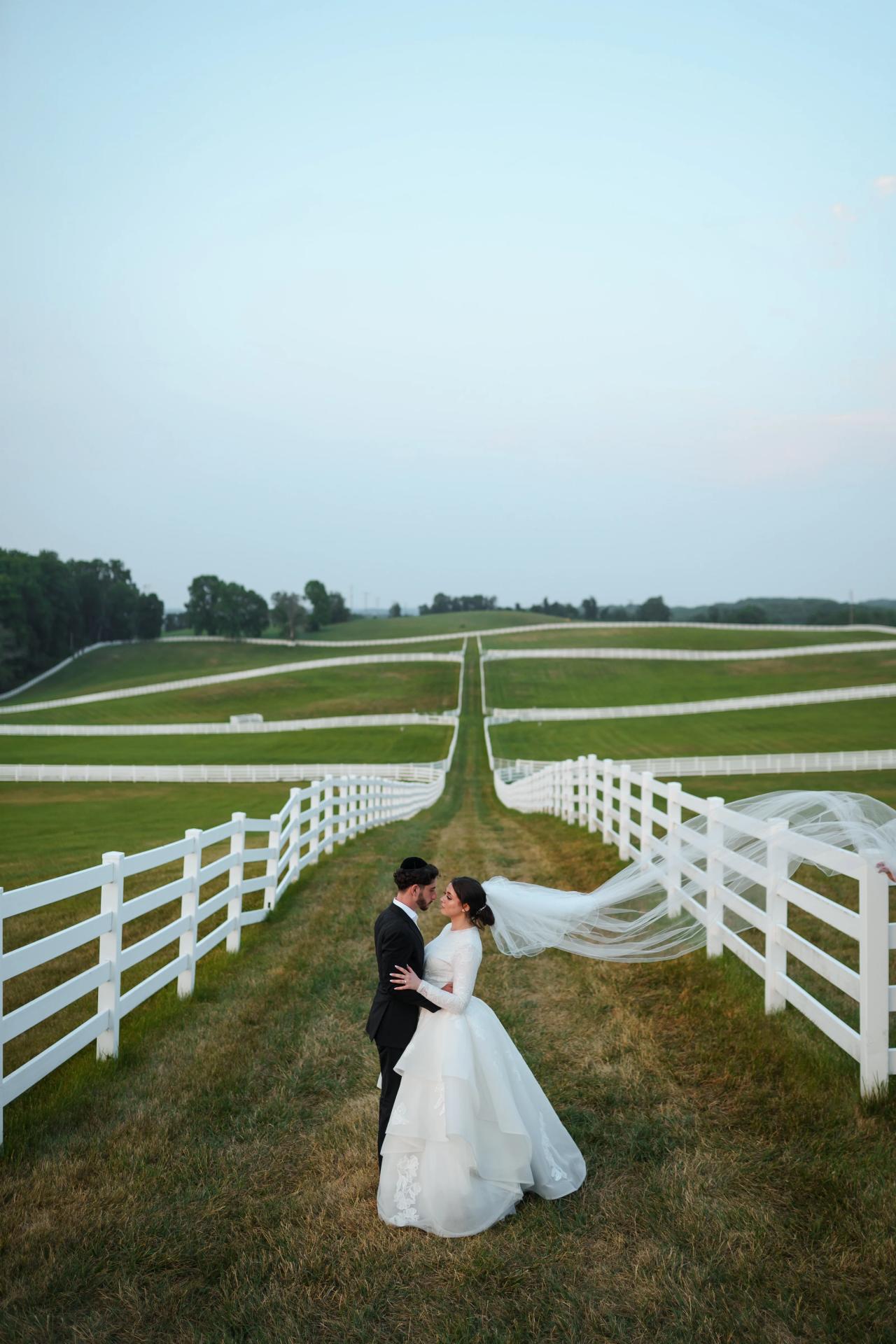 Bride and groom embracing between white countryside fences at dusk, with a flowing veil and pale blue sky at their Baltimore wedding. Wedding photography at Vignon Manor Farm, Baltimore, MD.