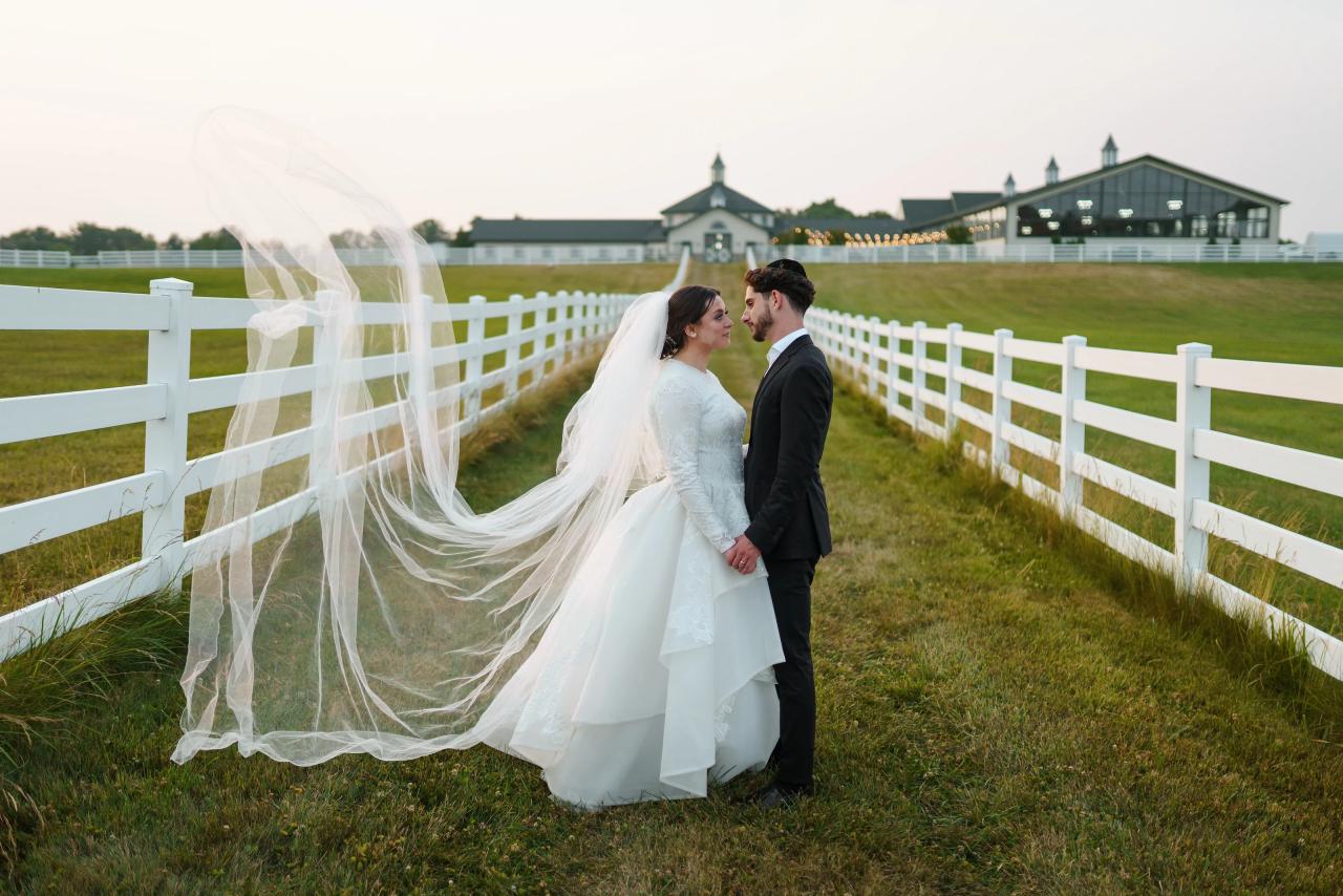 Bride and groom holding hands between white fences with the bridal veil lifted in the evening breeze at their Baltimore wedding. Wedding photography at Vignon Manor Farm, Baltimore, MD.
