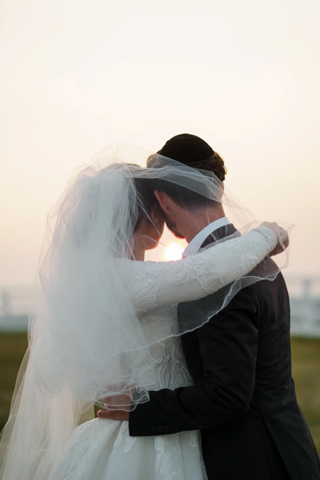 Bride and groom embracing beneath a translucent veil with warm sunset light shining between them at a Baltimore Jewish wedding. Wedding photography at Vignon Manor Farm, Baltimore, MD.