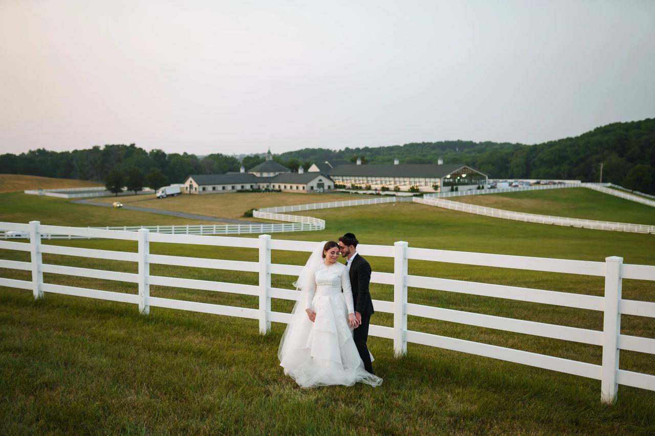 Wide dusk wedding portrait of the bride and groom beside white fences, rolling fields and an illuminated venue in Baltimore. Wedding photography at Vignon Manor Farm, Baltimore, MD.