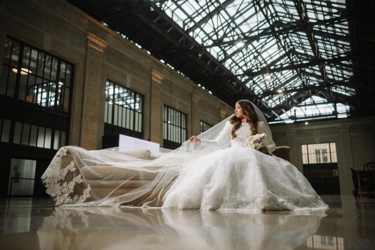 Bride in a white wedding gown walking with her veil flowing through a high-ceiling industrial hall, framed by arched glass roof and reflected on the polished floor.