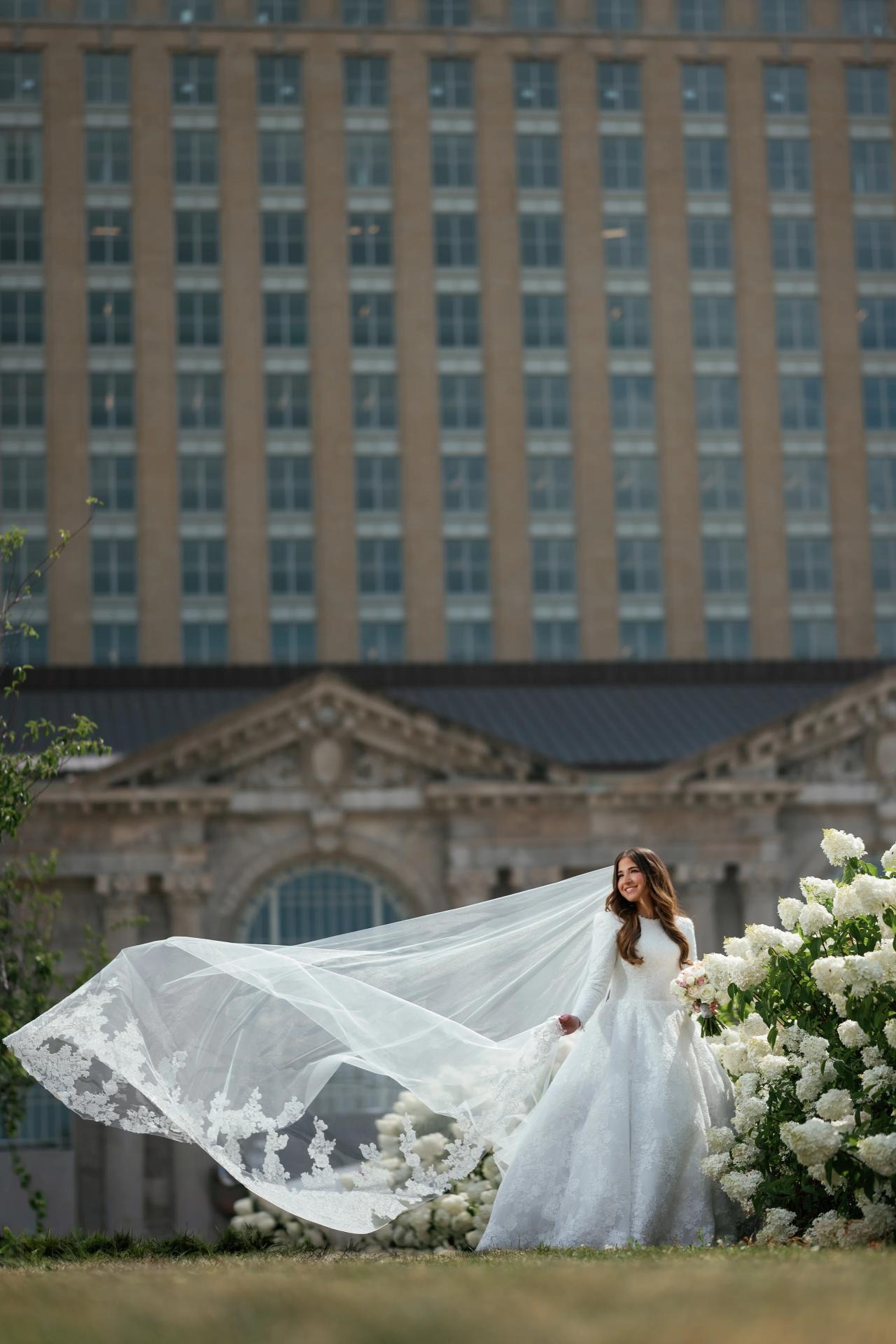 Bride in a white gown holding a bouquet with a flowing veil outdoors, framed by wedding florals and a large historic building with city skyline in the background.
