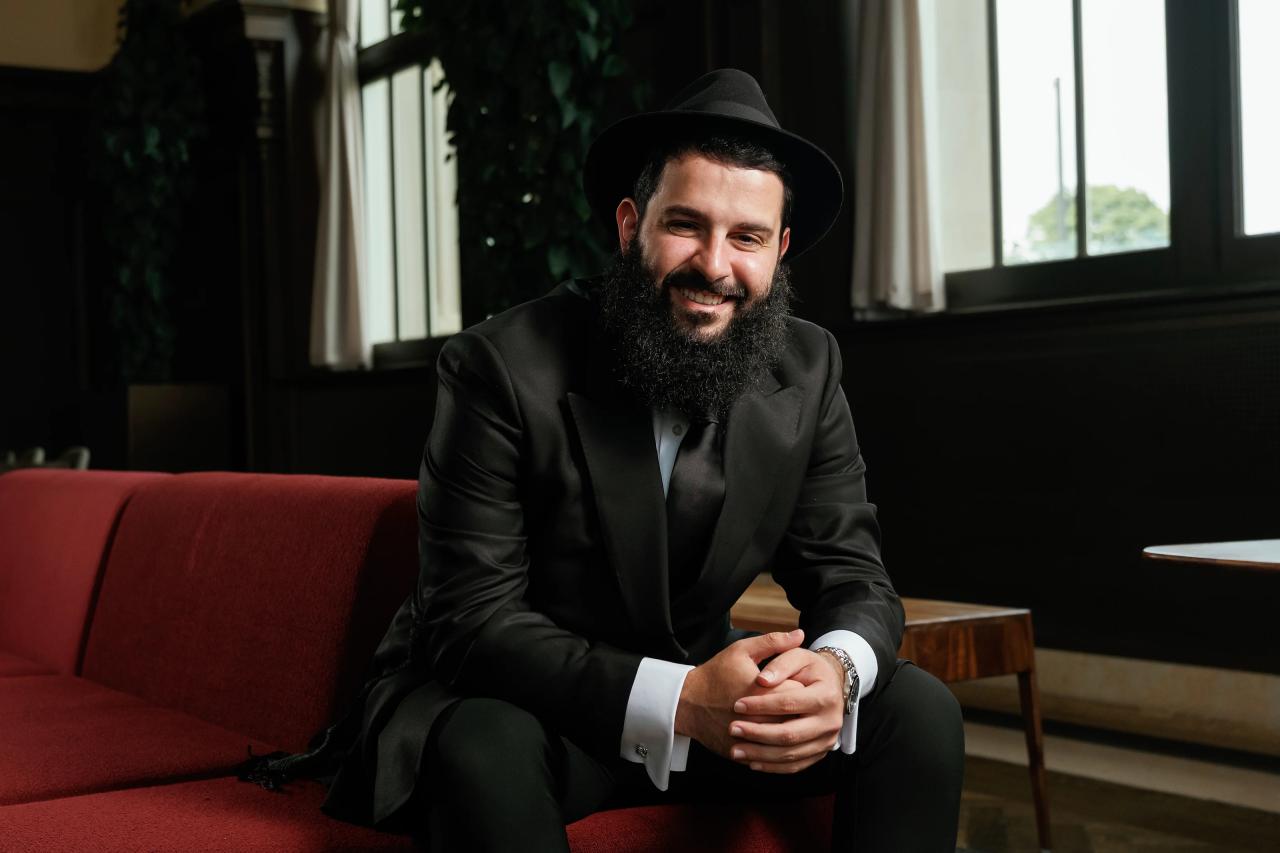 A groom in a black hat and suit seated on a red sofa, hands clasped, smiling softly in a dim lounge lit by a window.