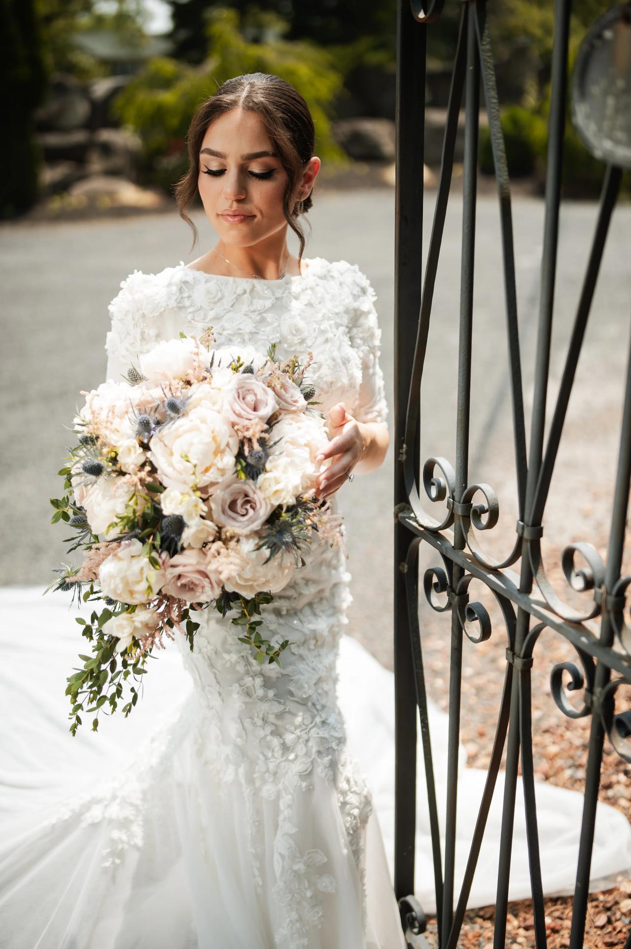 Framed portrait of a bride in a white lace gown holding a large bouquet beside a wrought-iron gate outdoors, softly lit with blurred greenery behind her. Wedding photography at The Barn At Indian Ridge, Highland, NY.