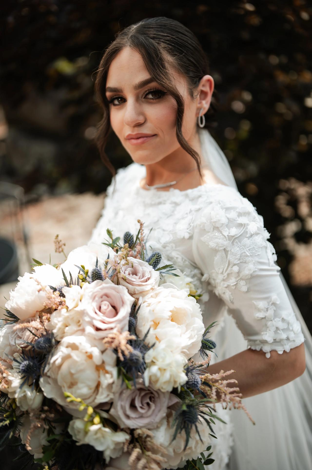 Close-up outdoor bridal portrait with the bride holding a lush bouquet, veil and white dress in warm golden light, with a dark blurred garden background. Wedding photography at The Barn At Indian Ridge, Highland, NY.