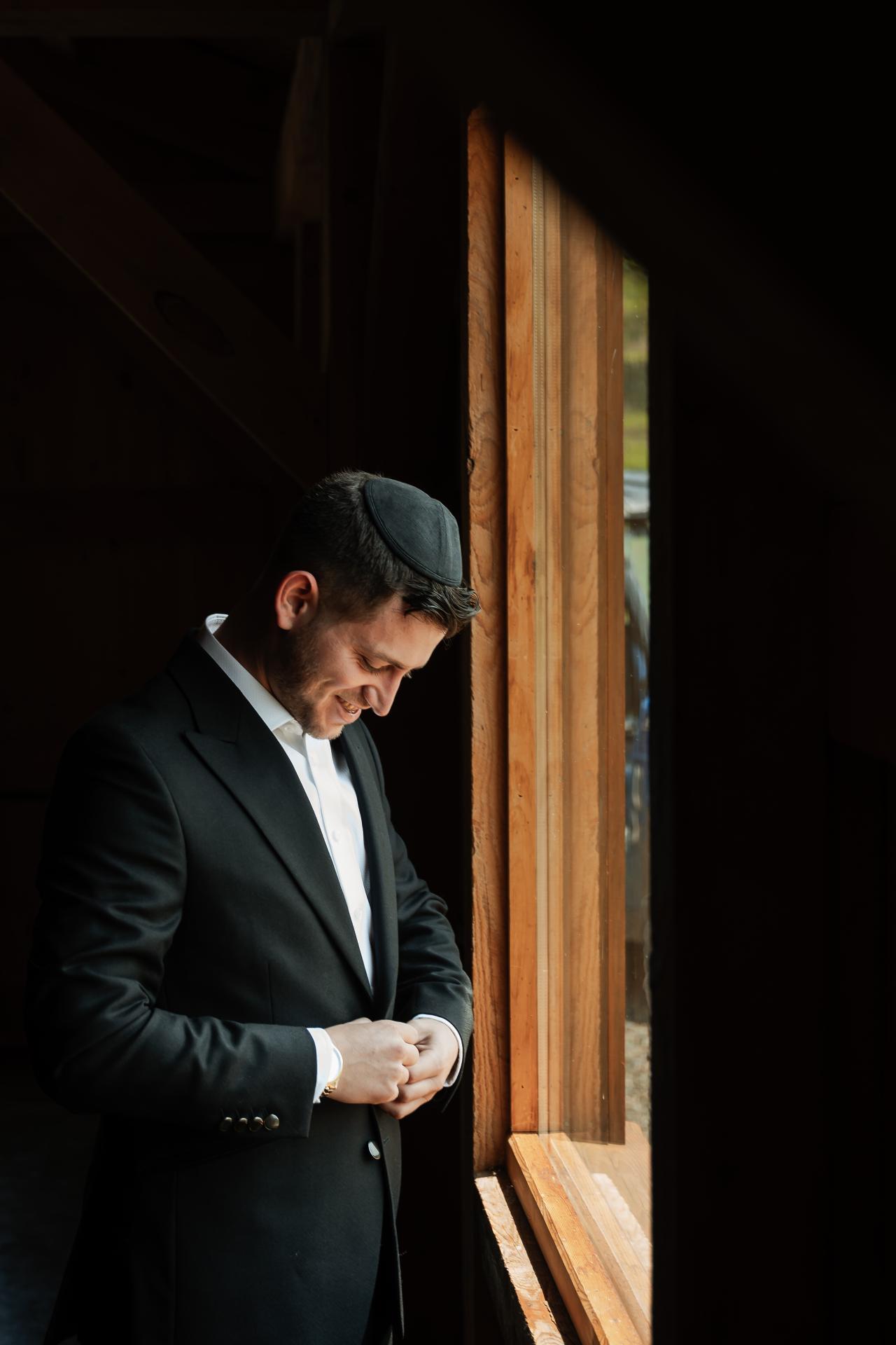 Groom in a suit adjusting tefillin in a warm beam of light near a wooden wall, solemn and intimate moment. Wedding photography at The Barn At Indian Ridge, Highland, NY.