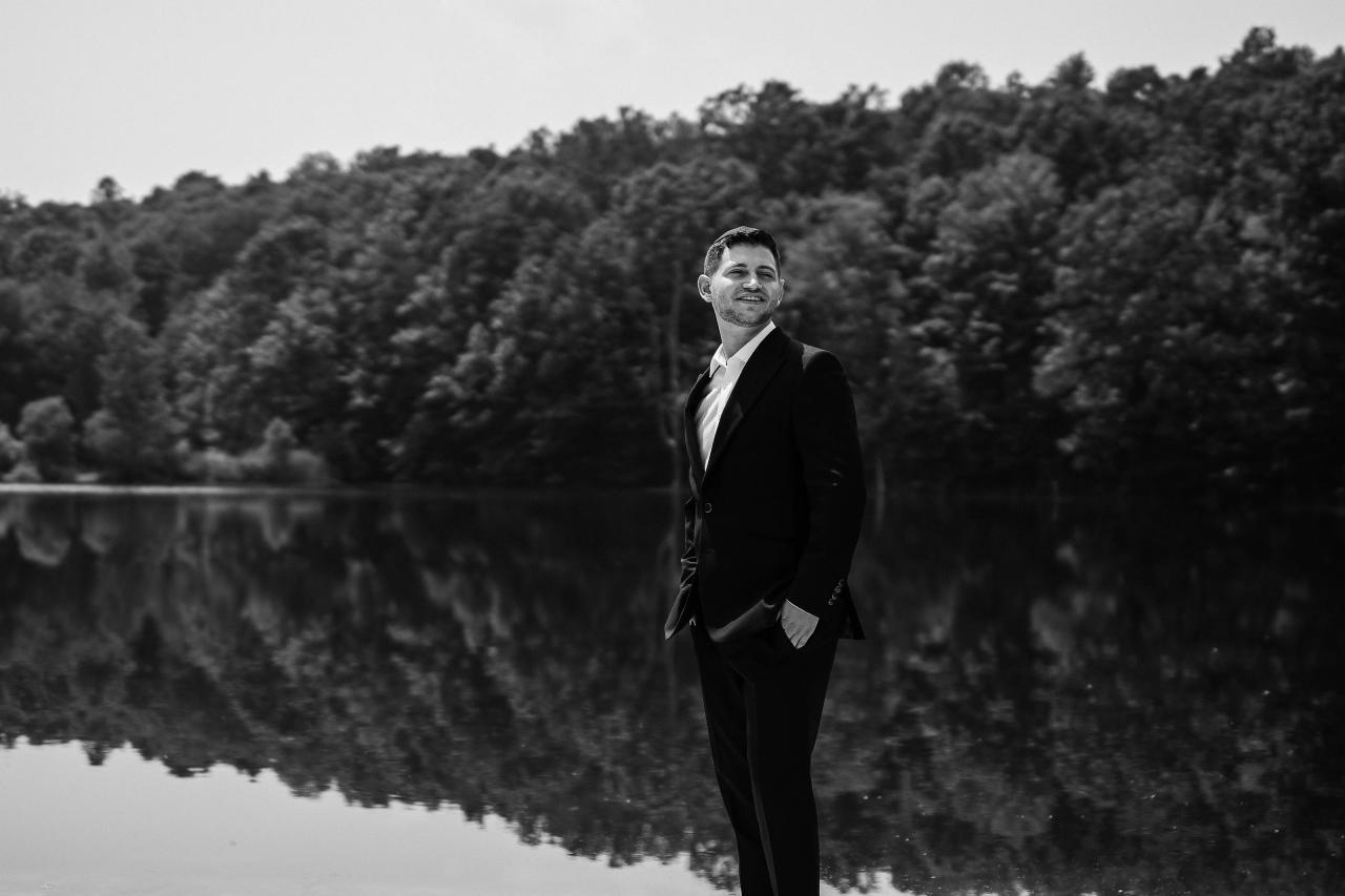 Black-and-white portrait of a groom standing by a calm lake with dense trees in the background, suit jacket and white shirt, smiling toward the camera. Wedding photography at The Barn At Indian Ridge, Highland, NY.