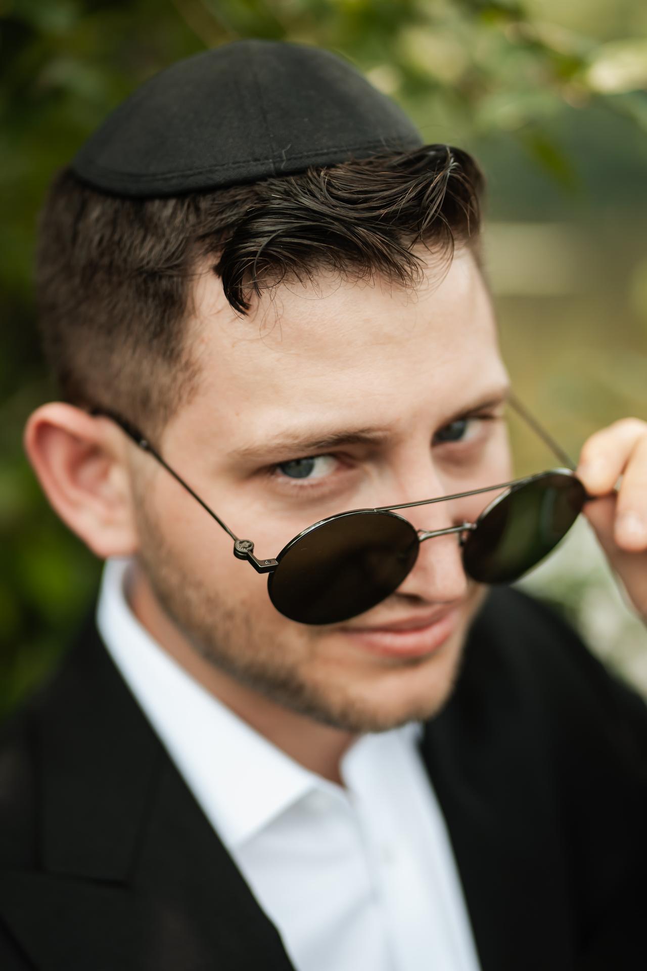 Outdoor close-up portrait of a groom wearing a black kippah and round sunglasses, looking sideways with a relaxed smile, with blurred greenery background. Wedding photography at The Barn At Indian Ridge, Highland, NY.