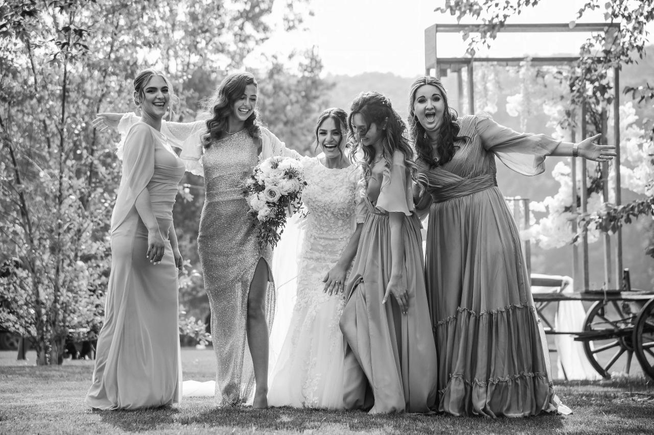 Black-and-white outdoor simcha photo of a bride in the center surrounded by joyful women in formal dresses, smiling and gesturing as they celebrate beside a pergola and garden greenery. Wedding photography at The Barn At Indian Ridge, Highland, NY.