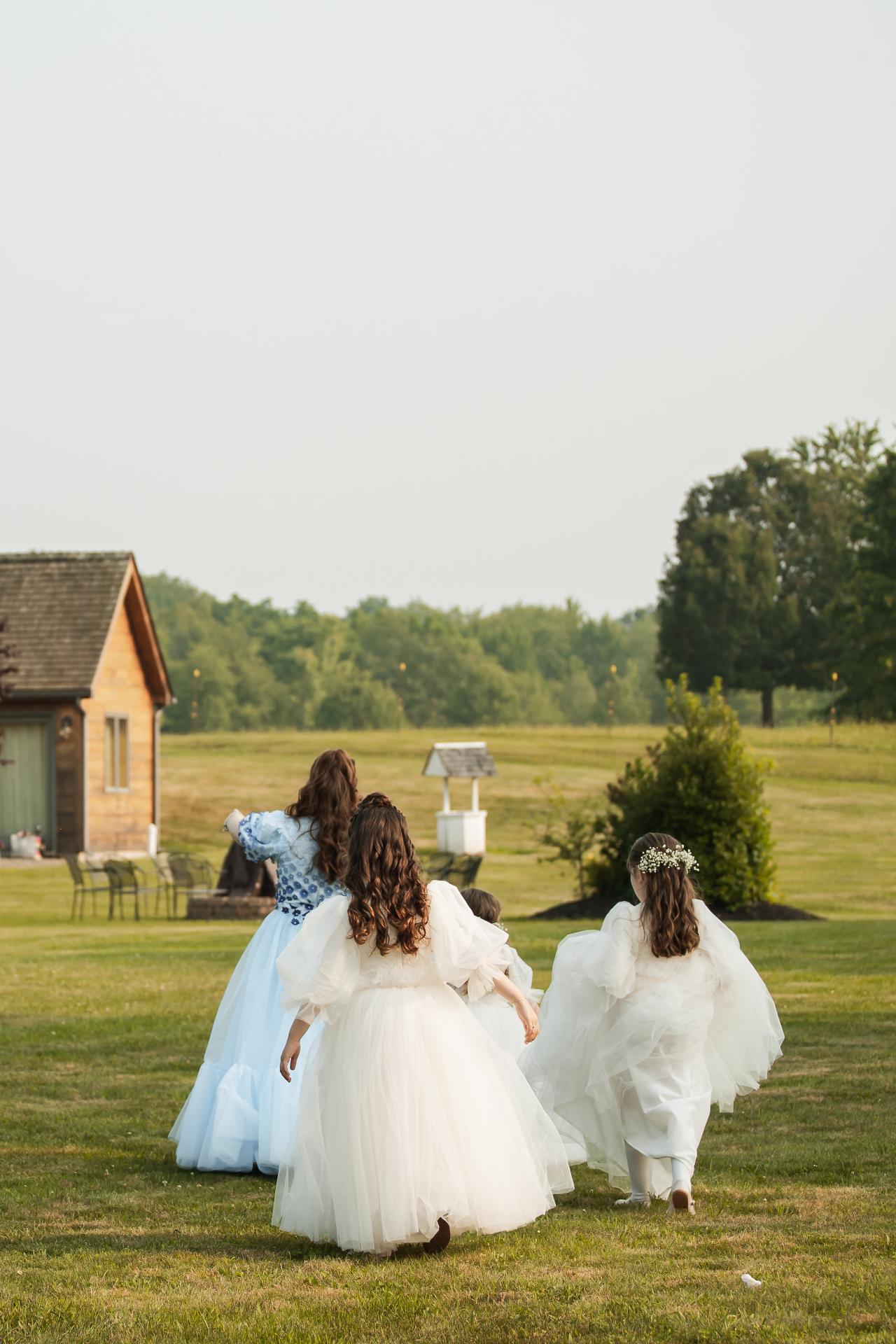 Three children in white dresses running outdoors on a grassy field at a simcha, seen from behind with open greenery in the background. Wedding photography at The Barn At Indian Ridge, Highland, NY.