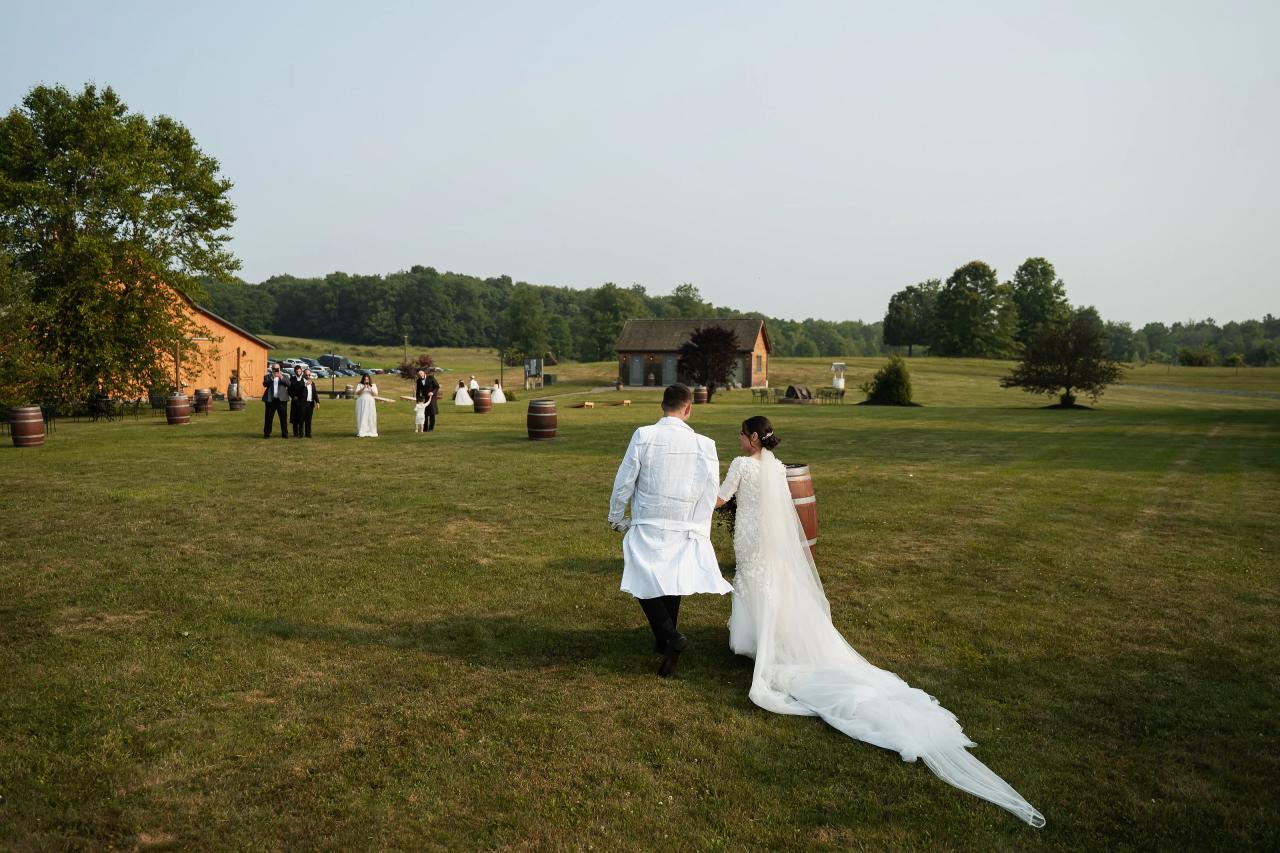Wedding couple walking hand-in-hand on a grassy field at an outdoor simcha, bride’s veil trailing behind, with rustic buildings and barrels in the background. Wedding photography at The Barn At Indian Ridge, Highland, NY.