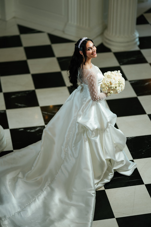 Bride in a white gown with a floral headpiece holding a white bouquet while walking and turning, photographed indoors on a black-and-white checkered floor.