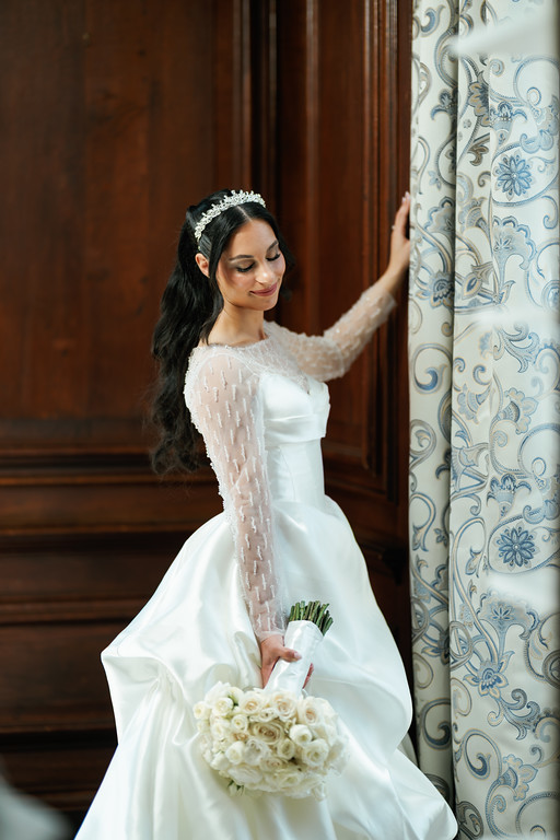 Frum bride in a white gown and lace sleeves stands by patterned curtains, holding a bouquet of white flowers and looking down with a serene expression.