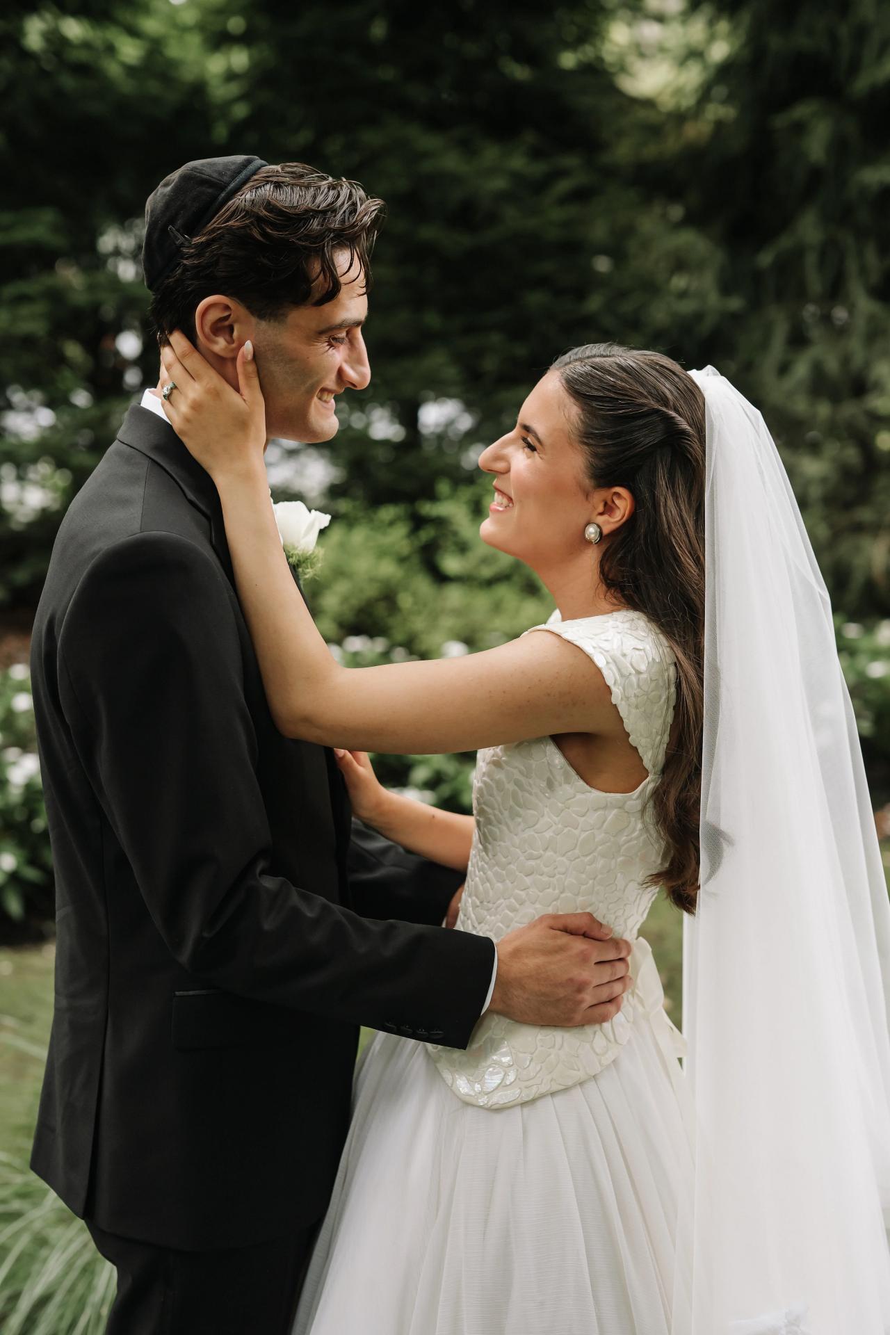 Groom and bride smiling at each other outdoors, groom adjusting the bride’s hair and veil while she rests her hand on his cheek; greenery background with soft bokeh. Wedding photography at Crystal Plaza, Livingston, NJ.