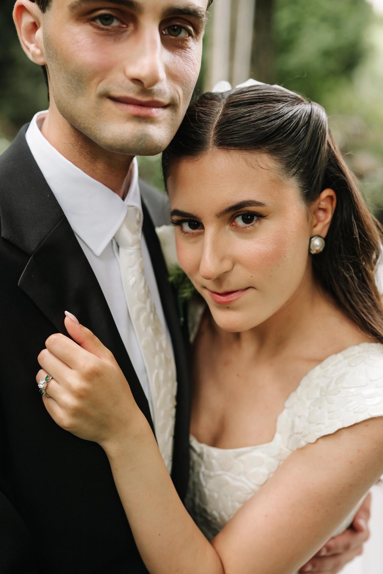 Close-up wedding portrait of a bride leaning into her groom outdoors, her hand on his lapel, with soft green background blur. Wedding photography at Crystal Plaza, Livingston, NJ.