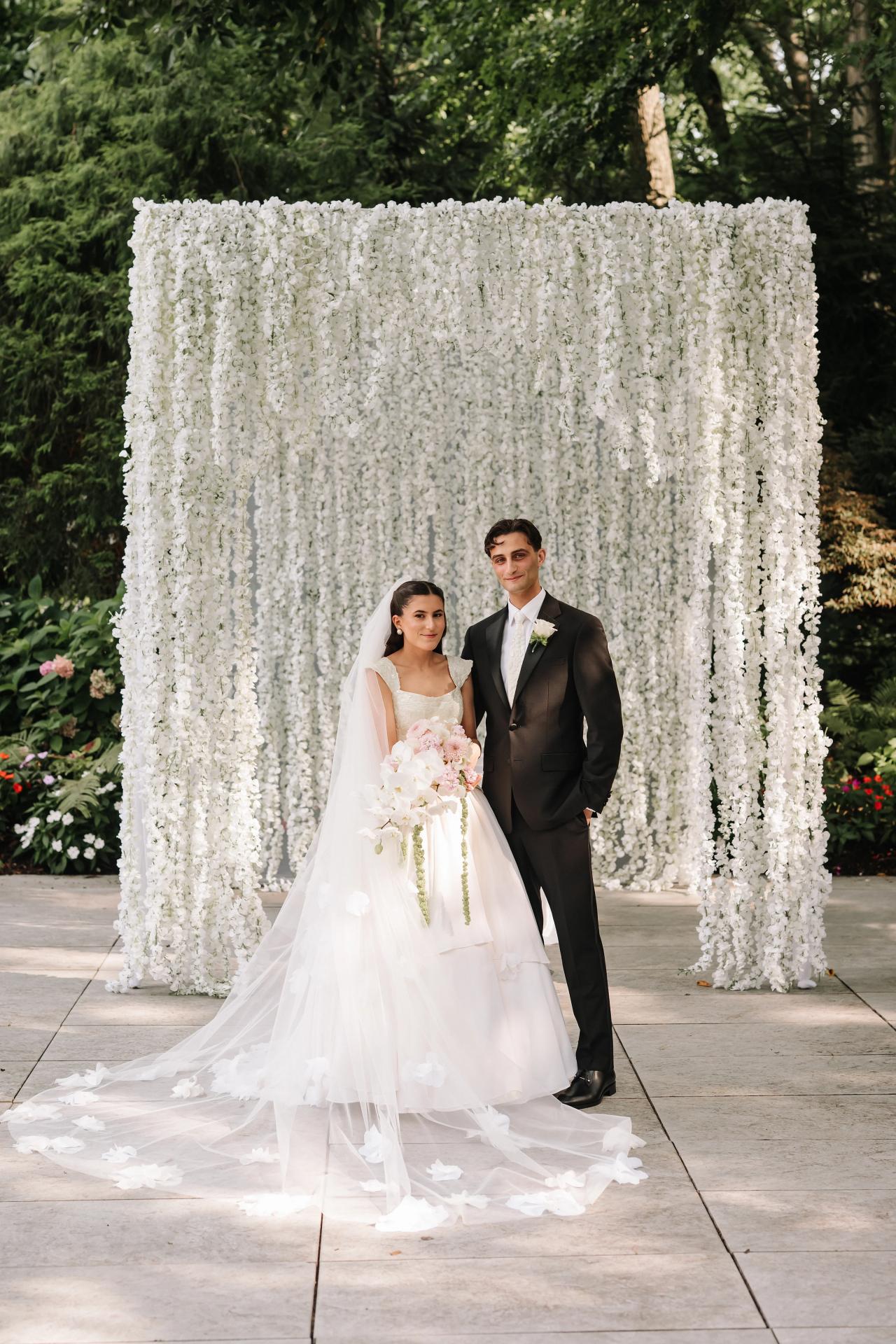Bride and groom standing beneath a white floral chuppah, bride holding a pink bouquet, outdoor ceremony portrait in bright natural light. Wedding photography at Crystal Plaza, Livingston, NJ.