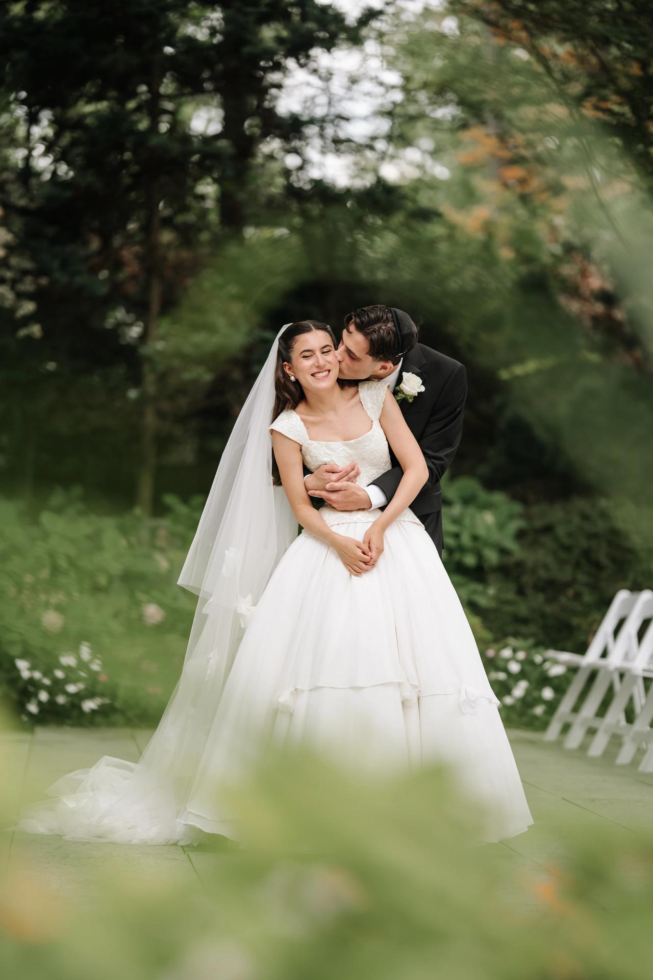 Bride and groom embracing outdoors in a garden, with the bride’s veil and soft foreground greenery creating a dreamy, blurred frame. Wedding photography at Crystal Plaza, Livingston, NJ.