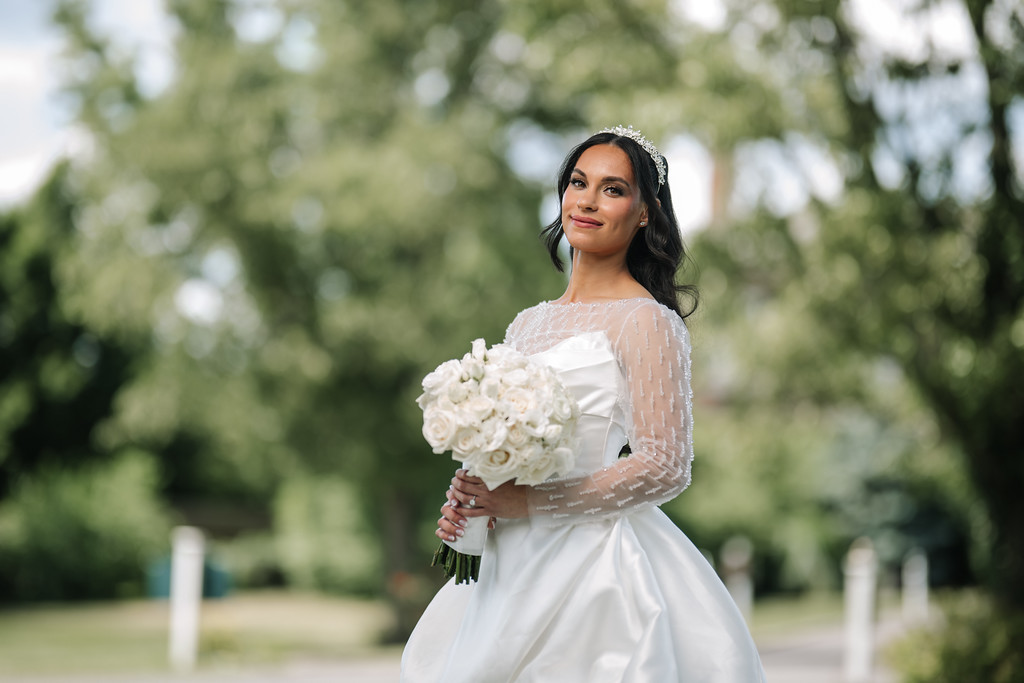 Bride in a white wedding dress with lace sleeves and a hair accessory holding a bouquet of white roses outdoors with greenery in the background.