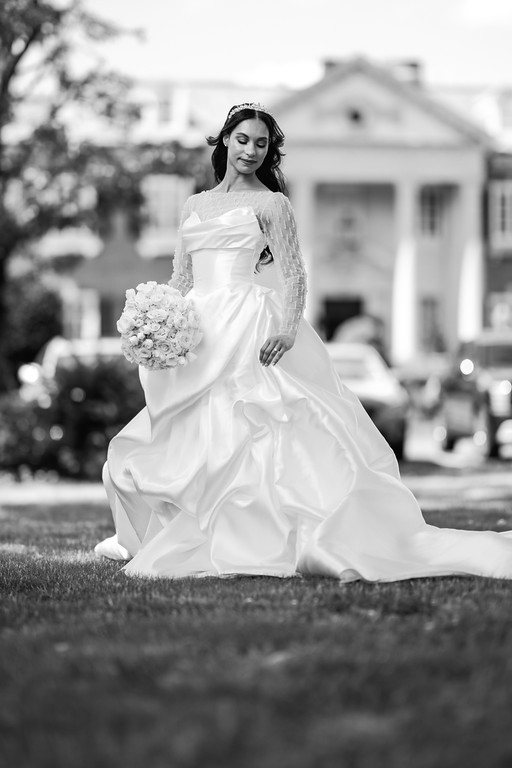Black-and-white portrait of a bride sitting on grass outdoors, holding a bouquet and smiling, with soft-focus buildings in the background.