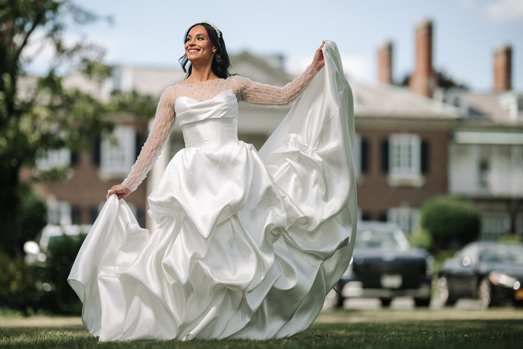 Smiling bride twirls outdoors in a white wedding dress, holding her skirt and veil in the breeze with buildings in the background.