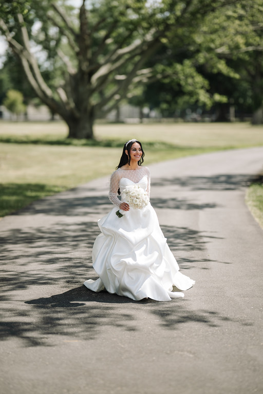 Bride in a white gown sitting on a sunlit park road, holding a bouquet and smiling at the camera.