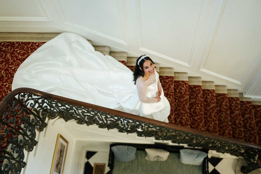 Smiling bride in a white dress and headpiece holding her pose on a staircase balustrade, looking back at the camera.