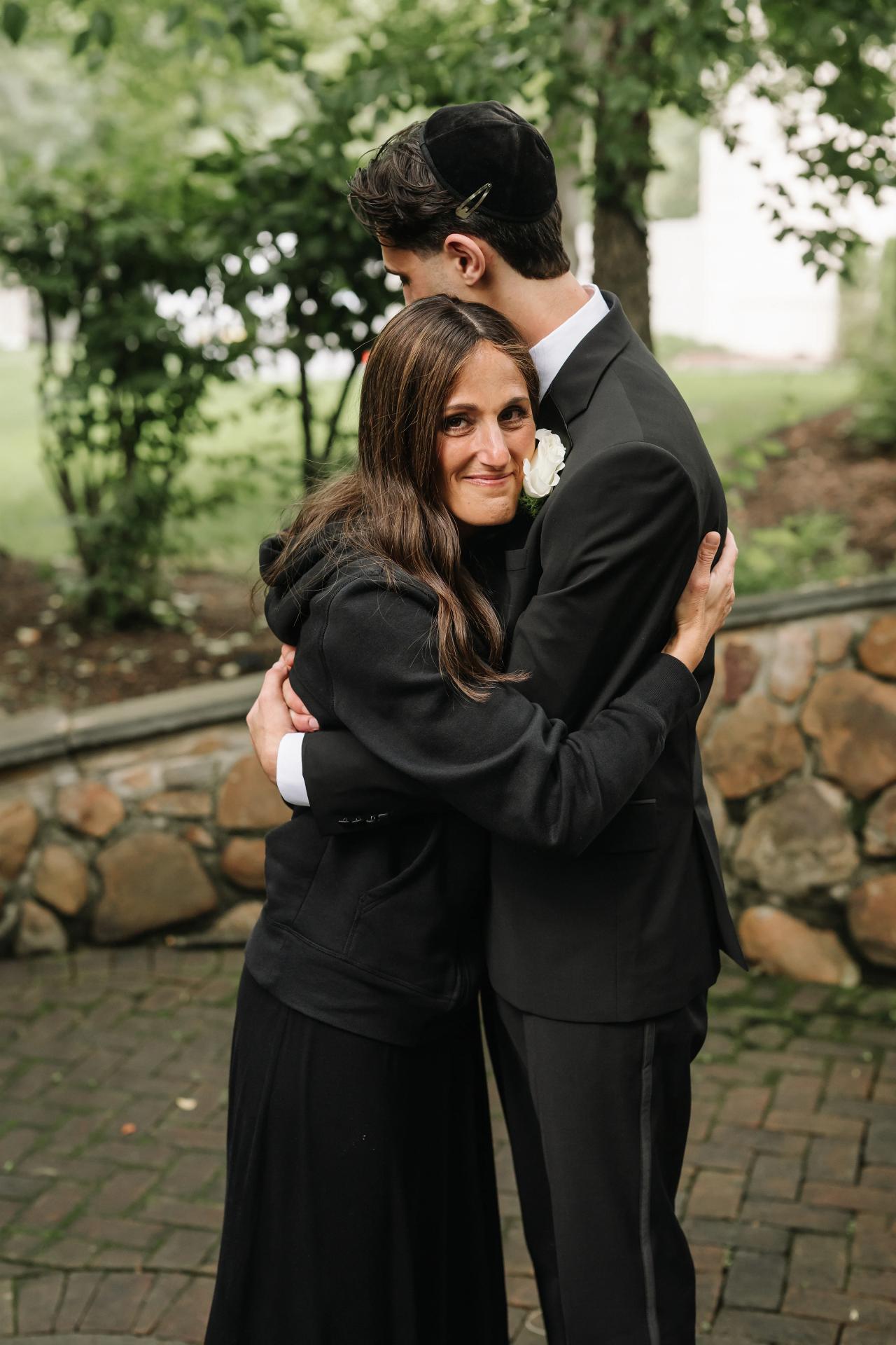 Framed hug between a bride and groom outdoors, smiling, with green trees softly blurred behind them and a stone wall and walkway in the foreground. Wedding photography at Crystal Plaza, Livingston, NJ.